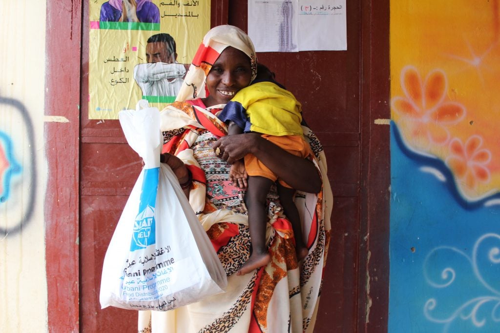 Smiling Sudanese woman holds child while showing relief package from Islamic Relief.